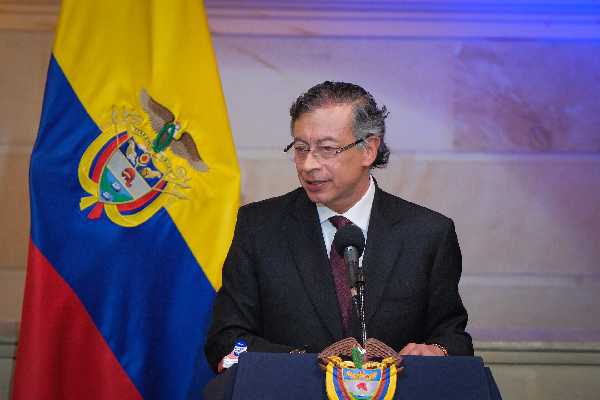 Gustavo Petro, wearing glasses and a dark suit, speaks at a lectern bearing Colombia's coat of arms, with the Colombian flag behind him