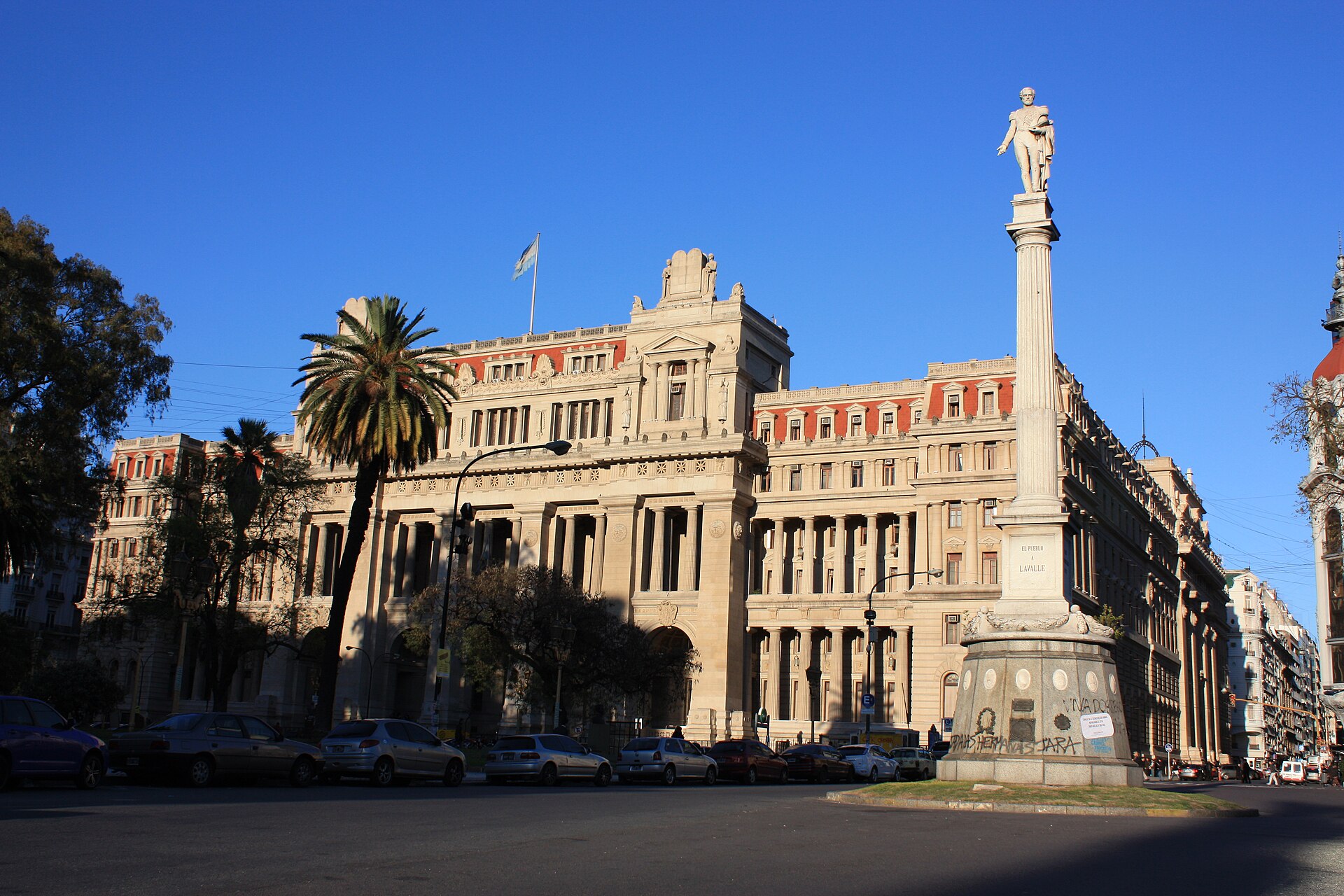 Beaux-Arts facade of the Palace of Justice in Buenos Aires with an Argentine flag on the roof, palm trees, parked cars and the Lavalle monument column in the foreground