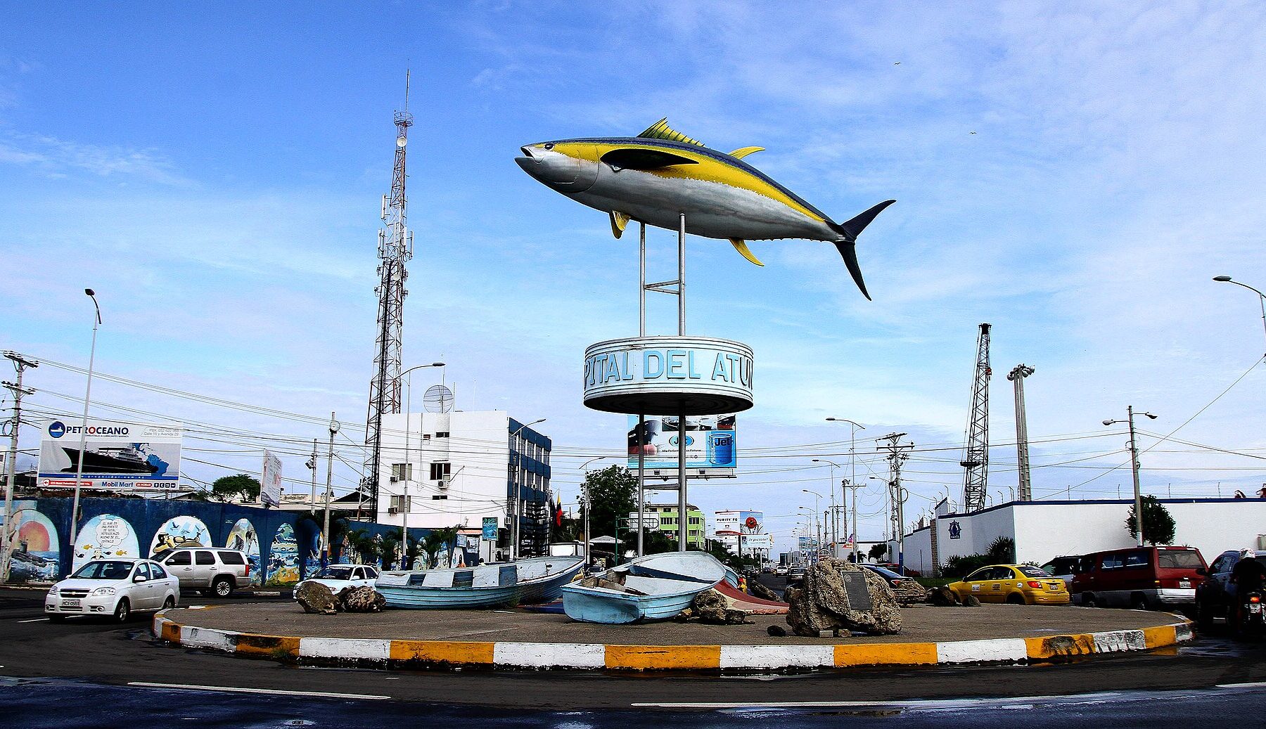 The tuna monument on a roundabout in Manta, Ecuador, with a Capital del Atún sign, small wooden fishing boats at its base and port cranes behind