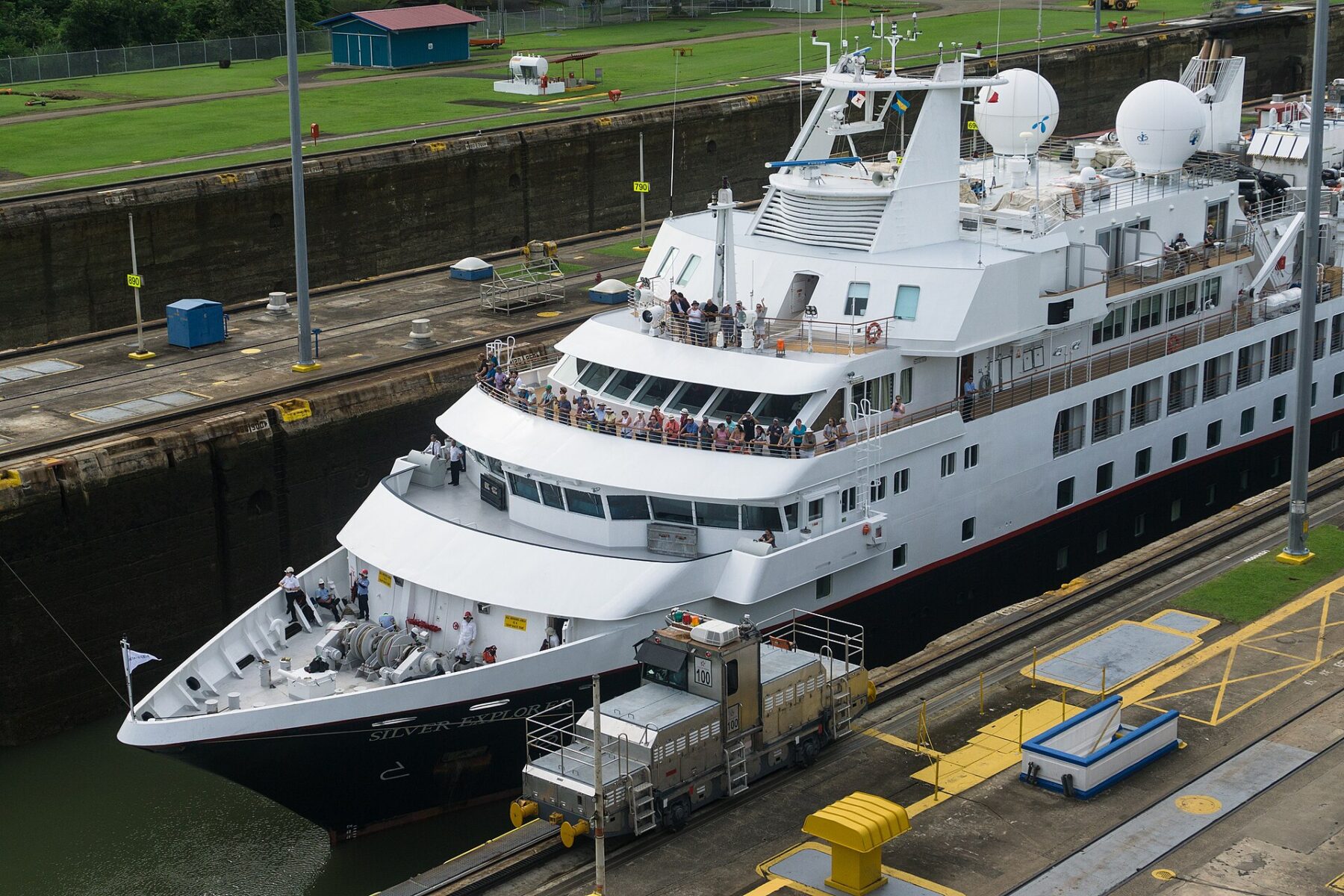 The small white expedition cruise ship Silver Explorer inside a Miraflores Locks chamber, passengers on its decks and a towing locomotive alongside