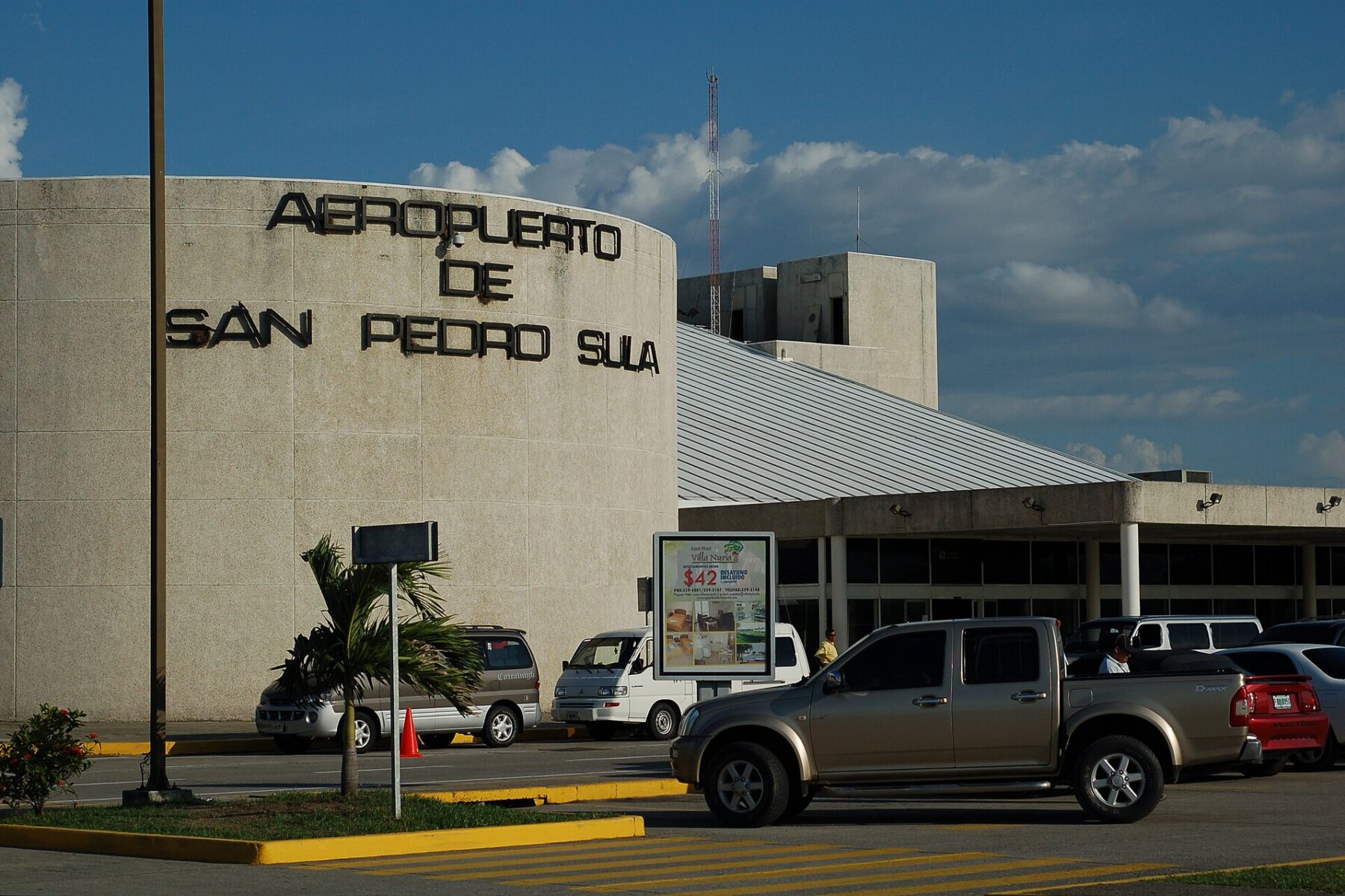 Curved concrete terminal building with the sign Aeropuerto de San Pedro Sula, pickup trucks and vans parked in front under a blue sky