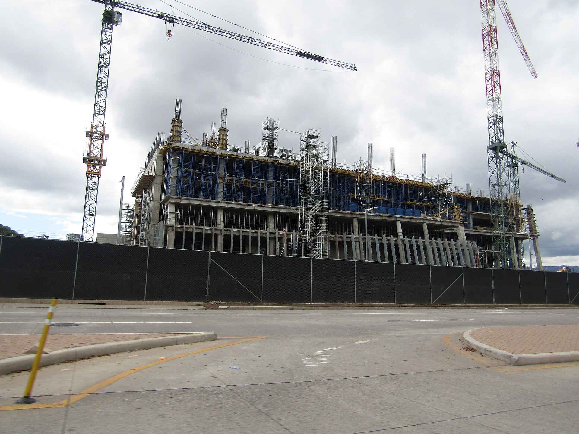 A multi-storey concrete building under construction with scaffolding and tower cranes behind a black fence beside an empty road