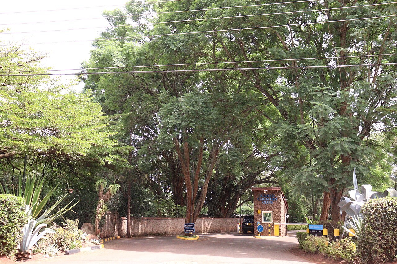 A guarded gate with a stone gatehouse and a Karen Country Club sign, set under tall green trees in Nairobi.