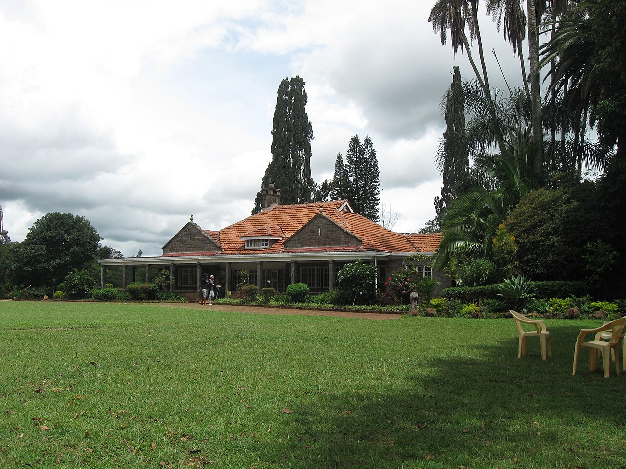 A low stone house with an orange tiled roof and a veranda behind a wide green lawn, surrounded by tall trees in Nairobi.