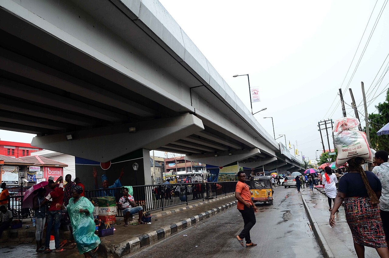 Pedestrians, street traders and a yellow tricycle taxi under a concrete flyover on a rainy day in Ikeja, Lagos.