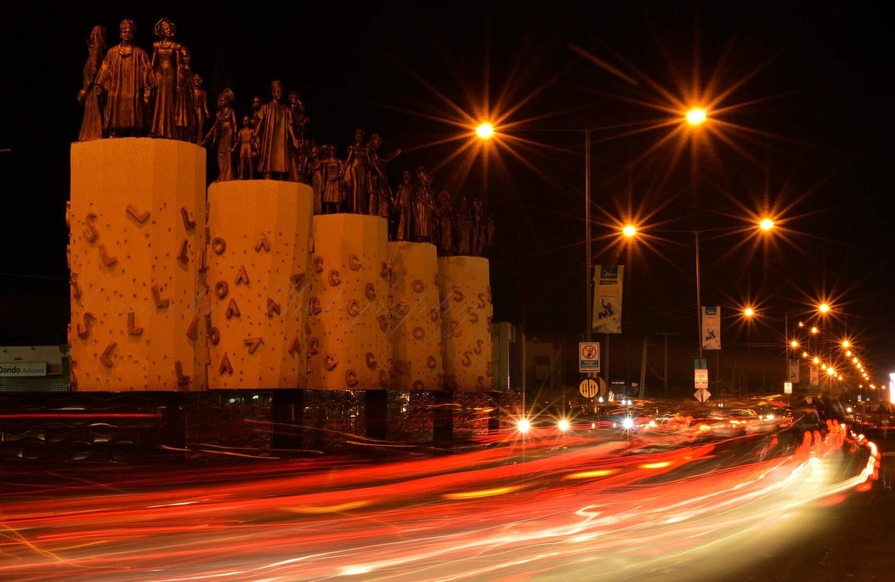 Bronze statues of men, women and children on tall yellow pillars beside a Lagos road at night, with streetlights and red streaks of car tail lights.