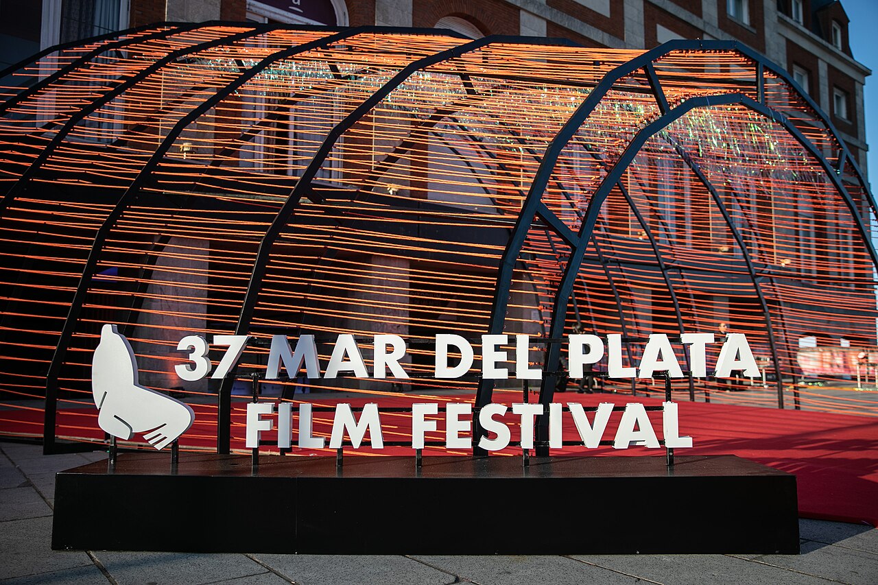 Sign reading 37 Mar del Plata Film Festival with a sea lion logo, in front of a red carpet and an orange-lit arch at a brick building