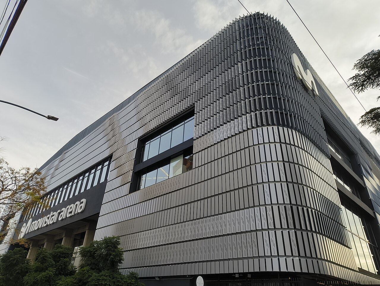 Curved corner of the Movistar Arena in Buenos Aires, clad in grey metal louvres, with the arena sign on the left and trees in front.