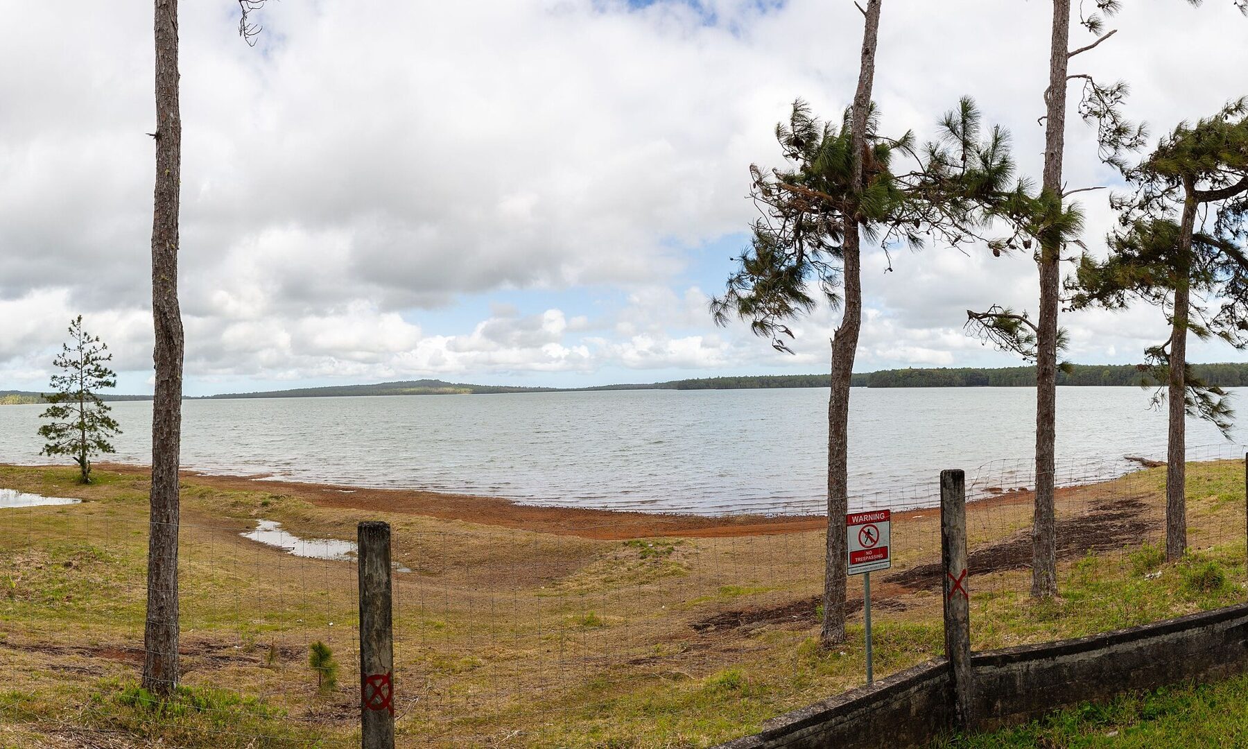 The Mare aux Vacoas reservoir in Mauritius with a red-brown exposed shoreline, pine trees, a fence and a no-trespassing sign under a cloudy sky