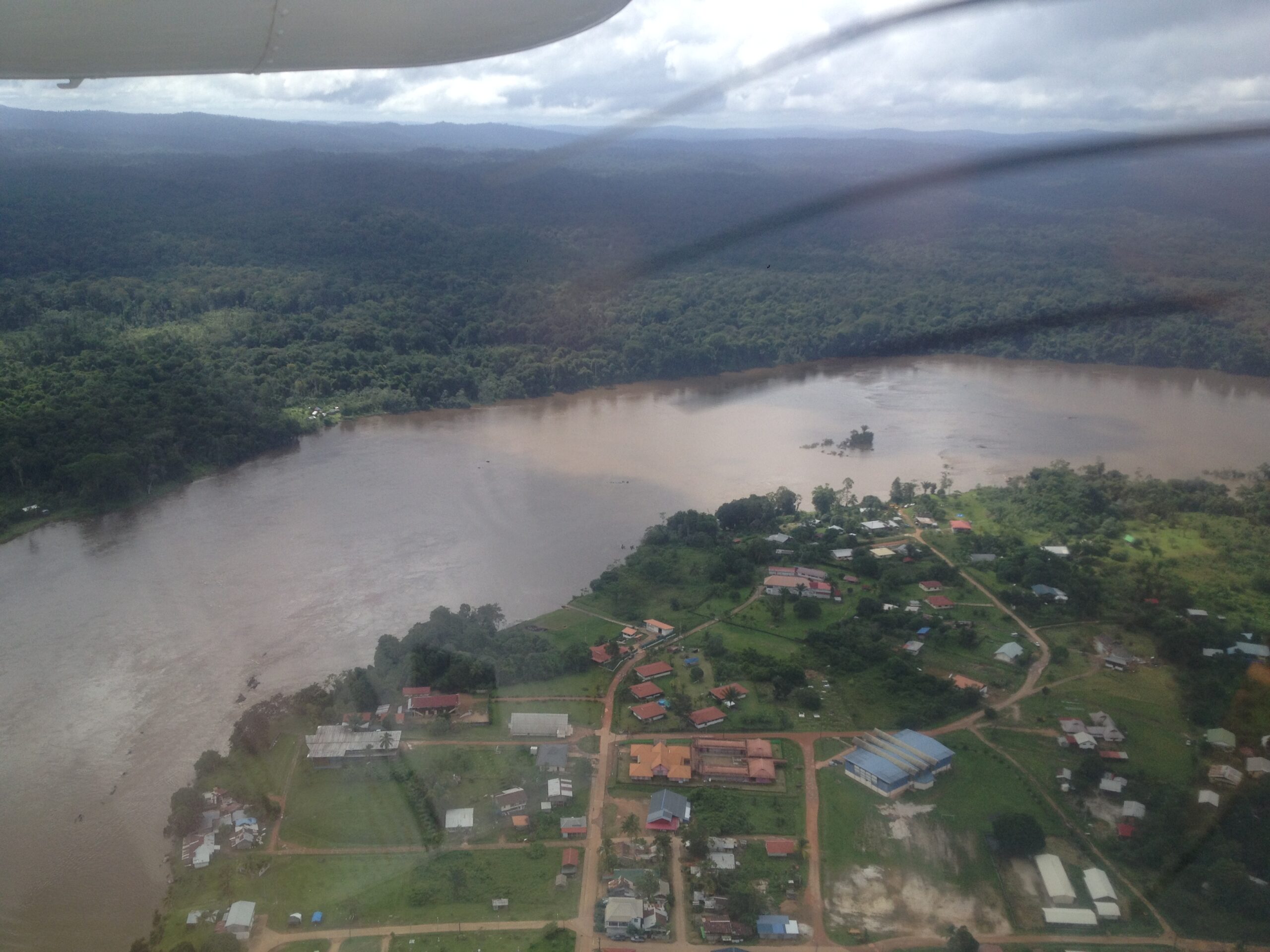 Aerial view of Maripasoula, a small town of red-roofed buildings on the bank of the wide, brown Lawa river, with dense forest beyond