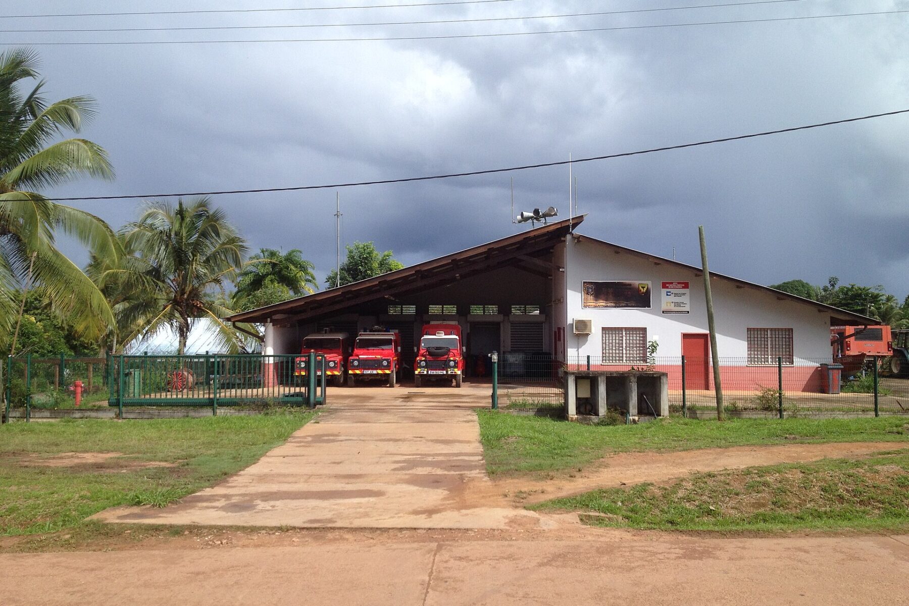 Three red fire engines parked in the open bays of a white and pink fire station with a sloping roof, palm trees and a red dirt road in front, under dark clouds