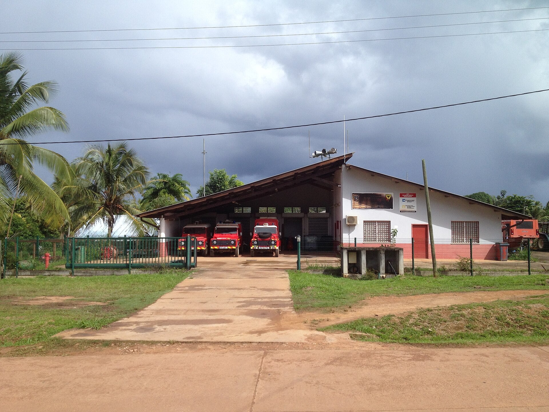 Three red fire engines parked in the open bays of a white and pink fire station with a sloping roof, palm trees and a red dirt road in front, under dark clouds