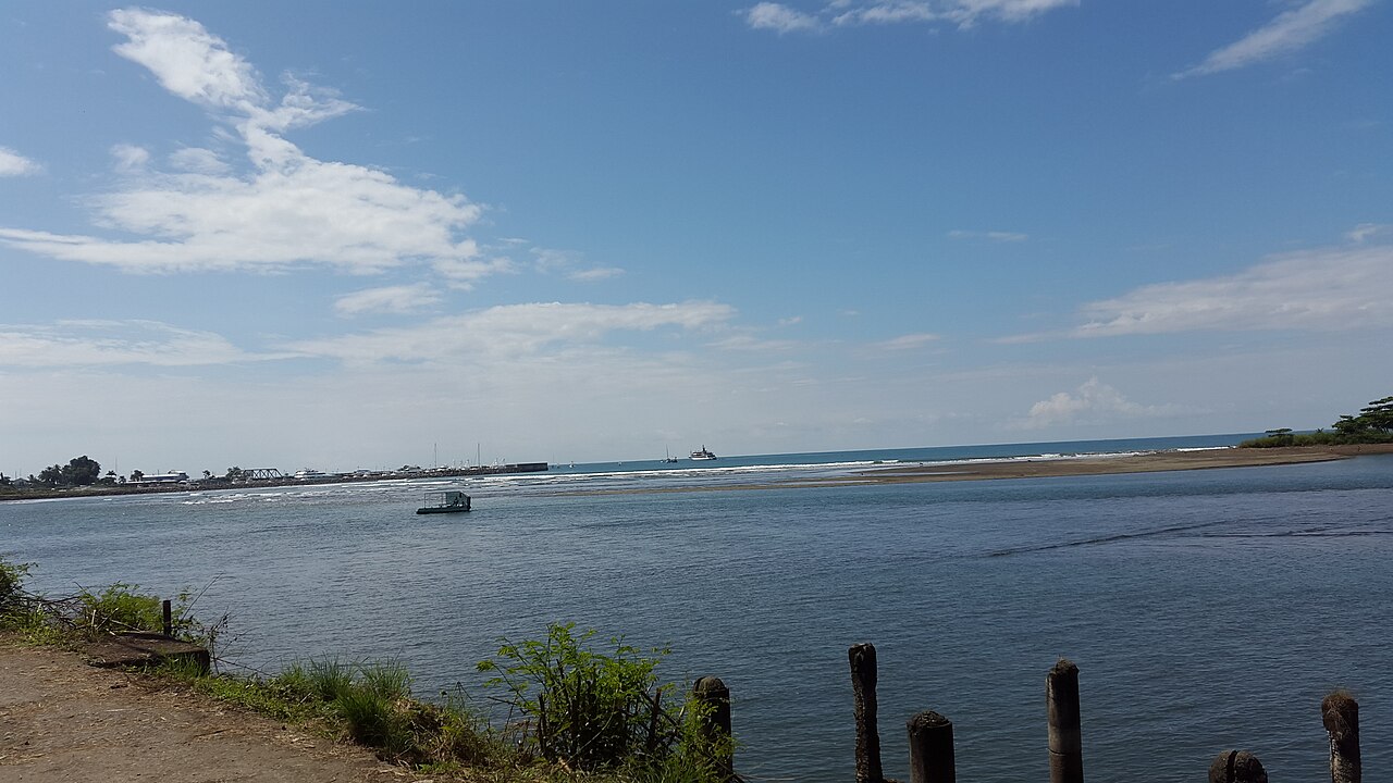 Calm water at the harbour mouth of Quepos, Costa Rica, with boats and a breakwater on the horizon under a blue sky.