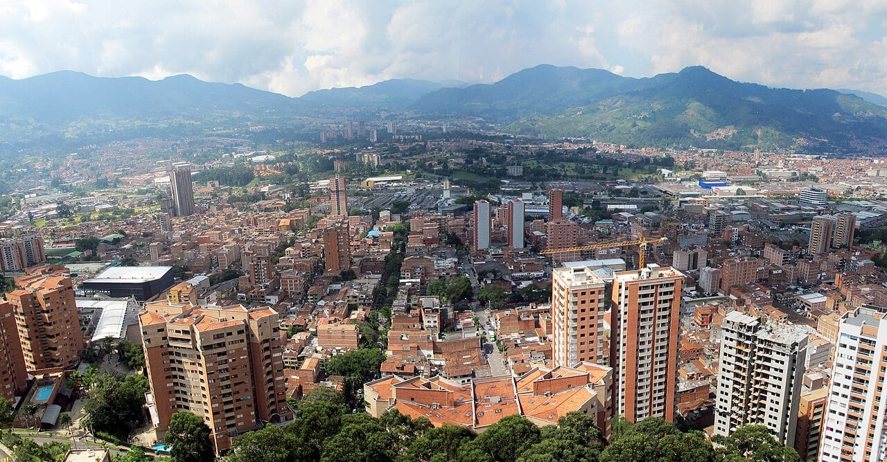Aerial view over Sabaneta, a town of brick apartment towers in the south of the Aburrá Valley, with green mountains and clouds behind.