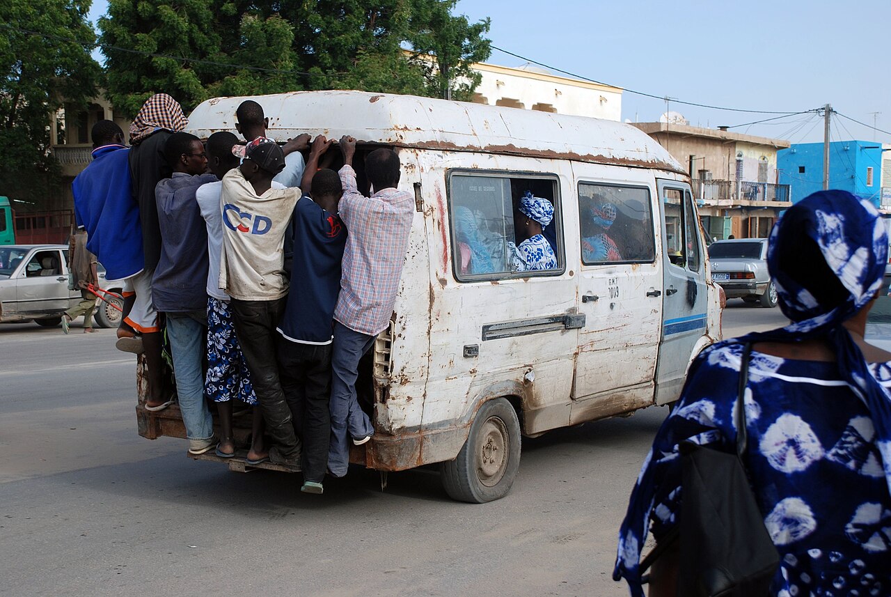 Passengers cling to the back of a crowded white minibus on a street in Nouakchott while a woman in a blue patterned wrap walks past