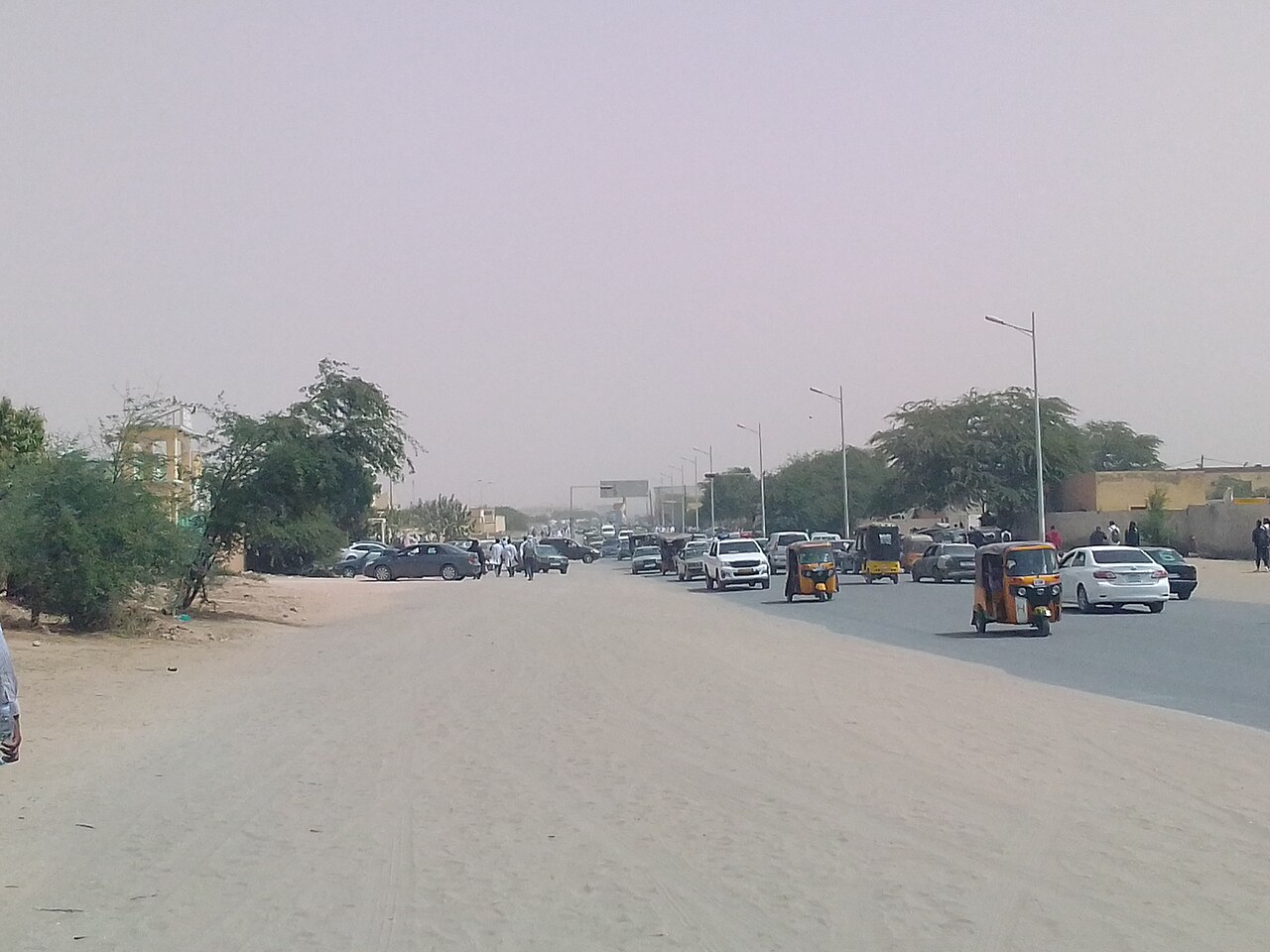 Cars and yellow three-wheeler taxis move along a sandy, dusty street in Nouakchott, Mauritania.