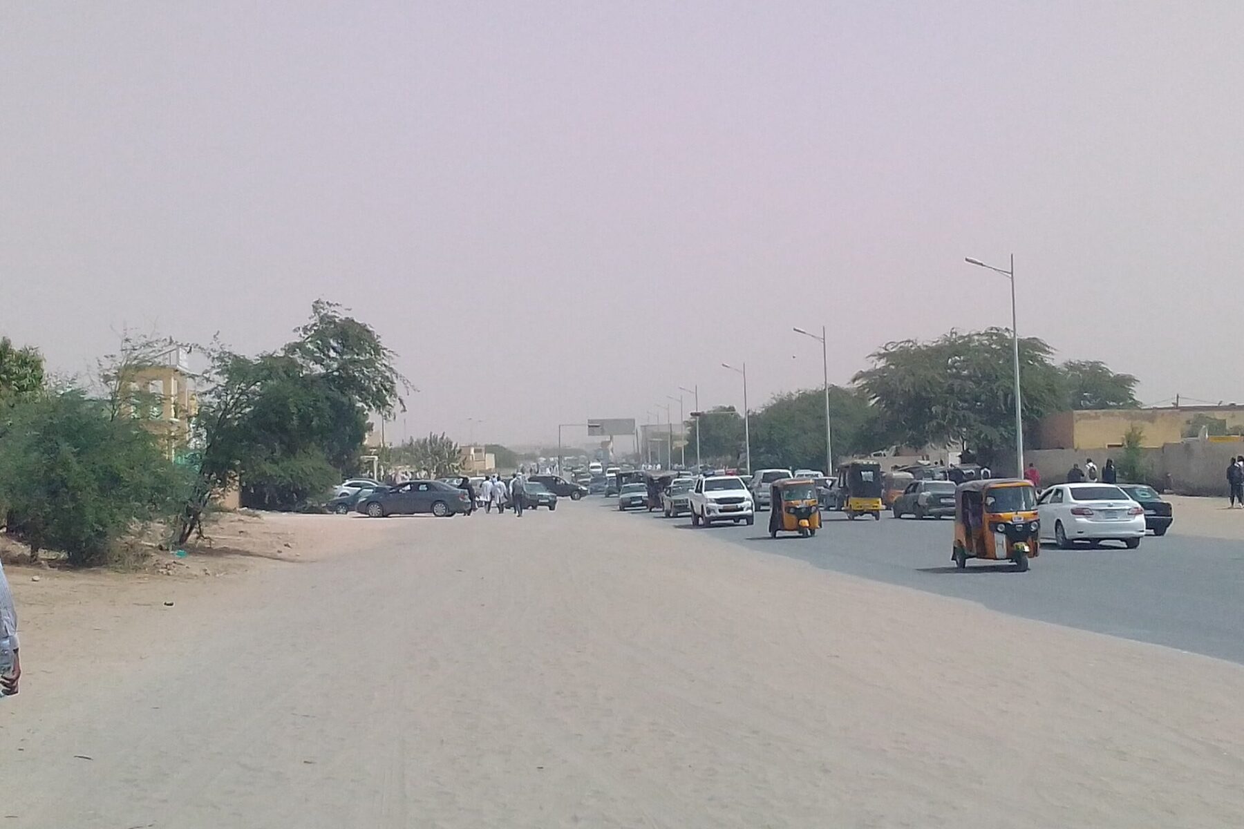 A wide road in Nouakchott, the capital of Mauritania, with cars and yellow auto-rickshaws under a hazy sky