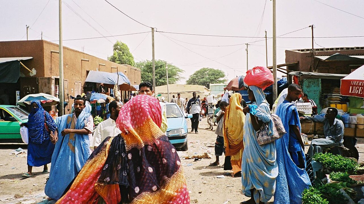 Shoppers in long robes and colourful wraps walk past vegetable stalls on a sandy market street in Nouakchott, Mauritania