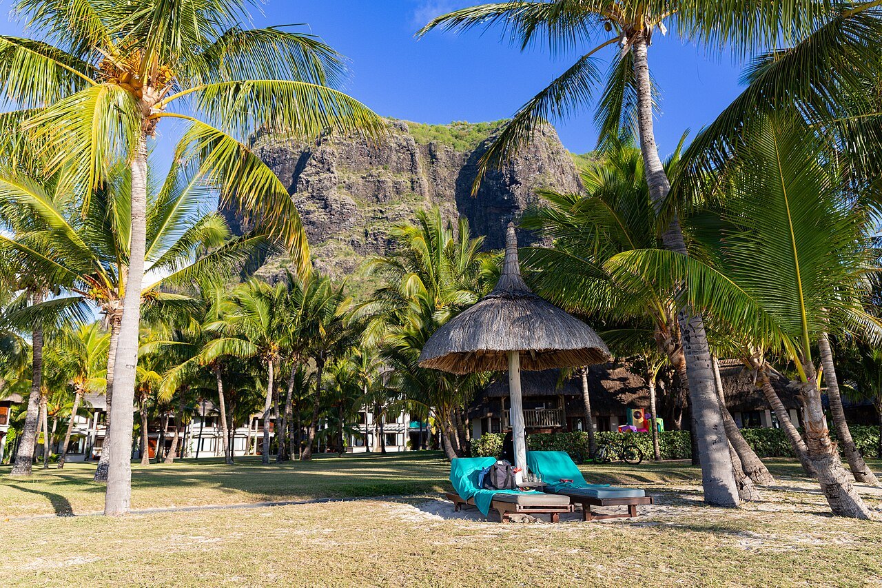 Sun loungers and a thatched umbrella under palm trees at a beach resort, with the rocky Le Morne Brabant mountain behind