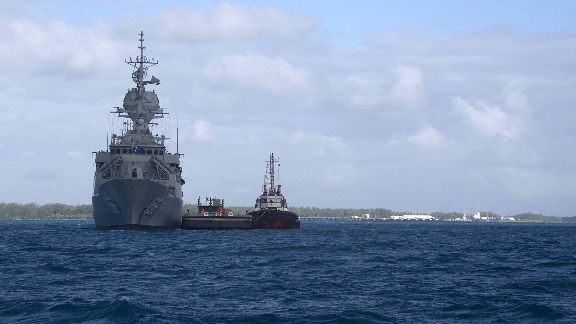 The Australian frigate HMAS Toowoomba at anchor off Diego Garcia with a tug and a barge, and the island pier and a container ship on the horizon