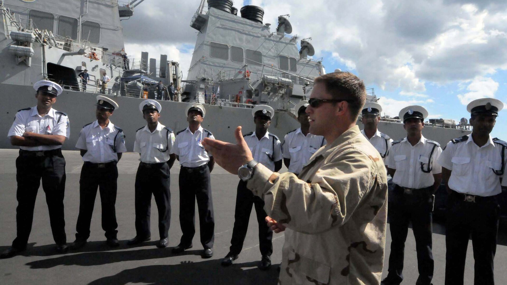 A US Navy sailor briefs uniformed Mauritian National Coast Guard sailors on a quay in Port Louis with a US destroyer behind them