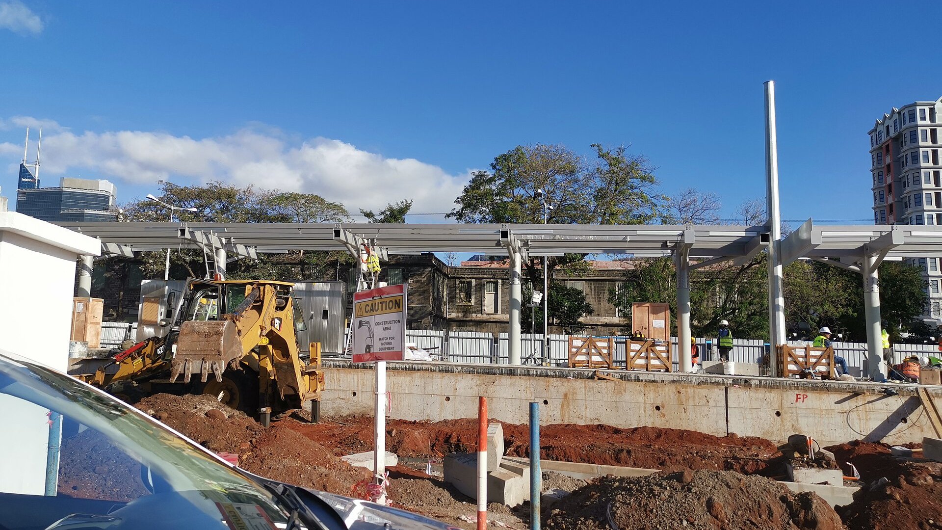 Construction site of a Metro Express light-rail station in Port Louis, Mauritius, with a backhoe and workers under a steel canopy