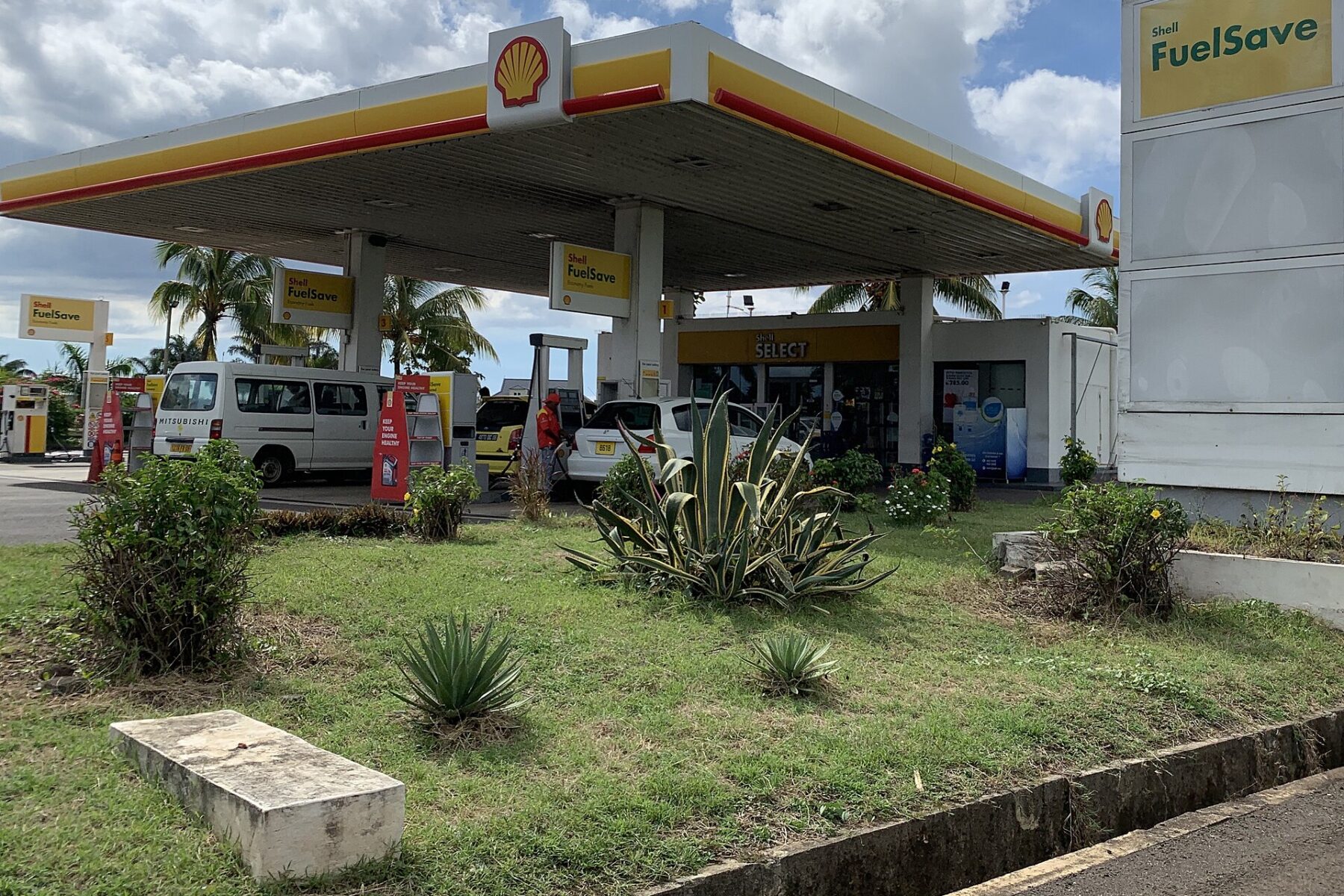 A Shell petrol station under a yellow and white canopy with palm trees behind it, a van and cars at the pumps, between Cascavelle and Bambous in western Mauritius