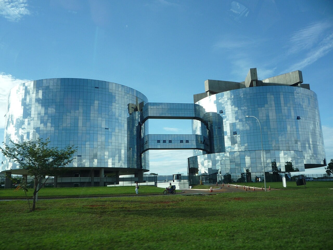 Two curved glass office towers joined by glass bridges under a blue sky, the Attorney General's Office headquarters in Brasília