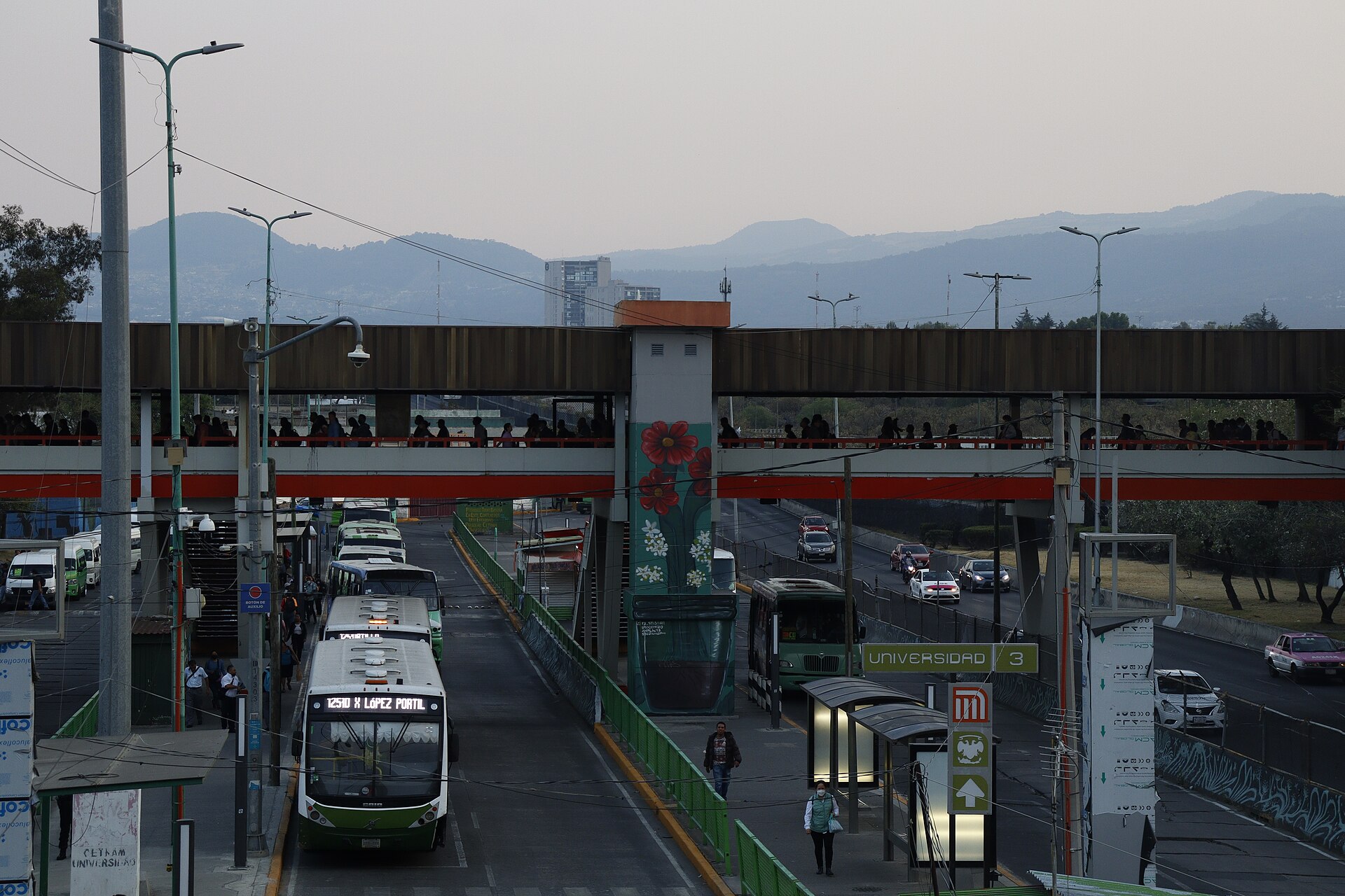 Green city buses lined up under an orange pedestrian footbridge at a Mexico City bus terminal, a Metro Universidad Line 3 sign and hills behind