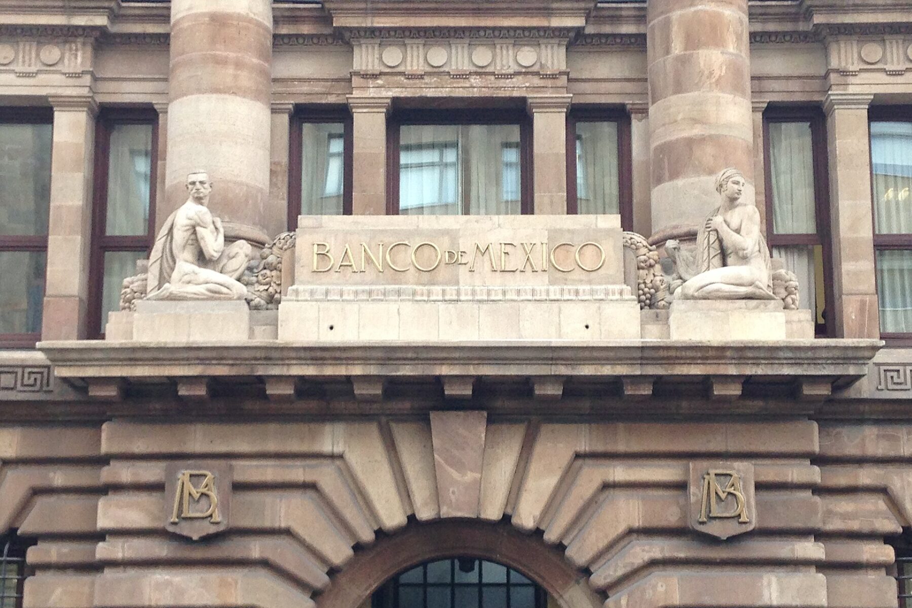Stone facade of the Banco de México (Mexico's central bank) building in Mexico City, with the bank's name and two sculpted figures above the entrance arch
