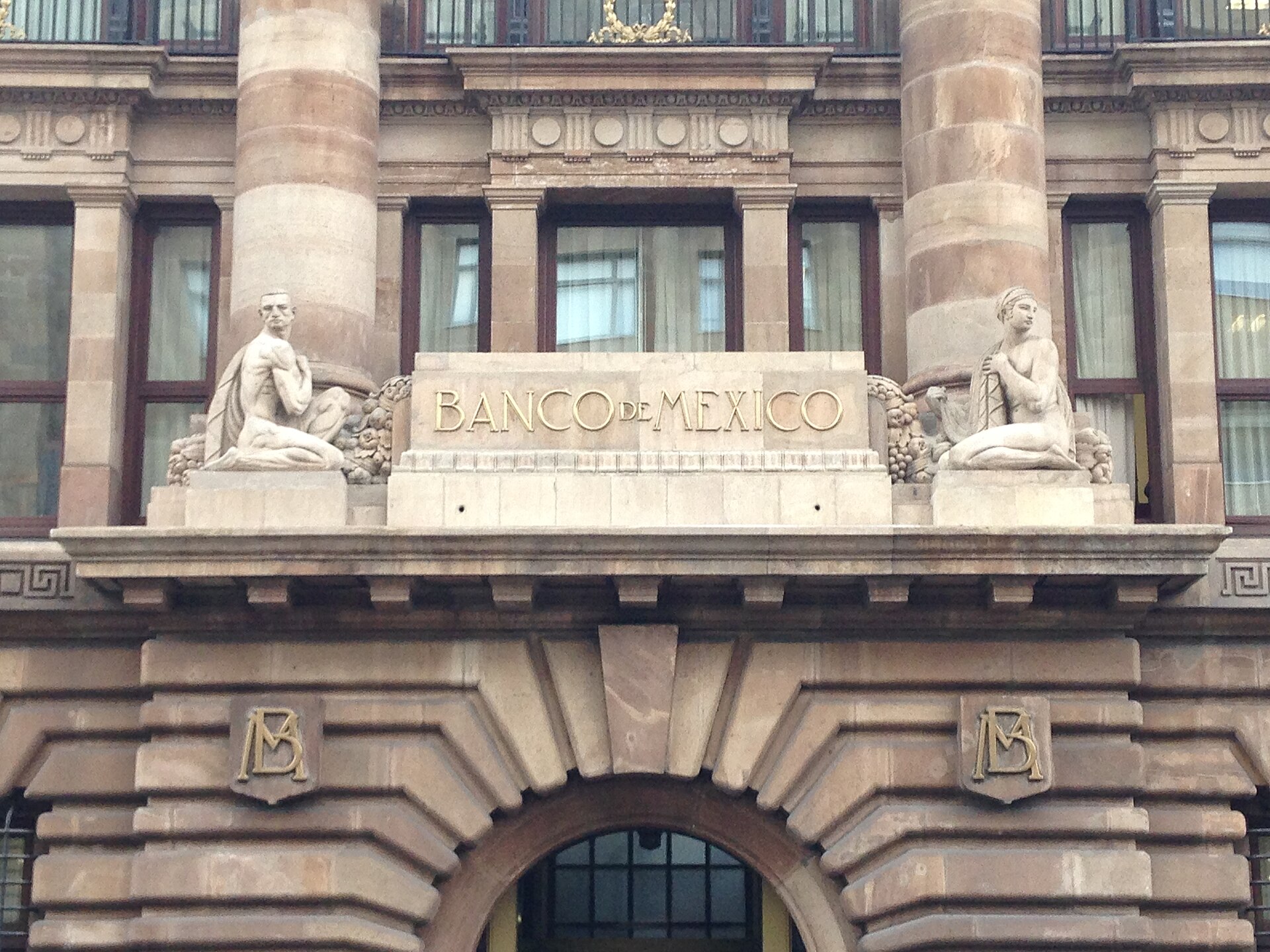 Stone facade of the Banco de México (Mexico's central bank) building in Mexico City, with the bank's name and two sculpted figures above the entrance arch