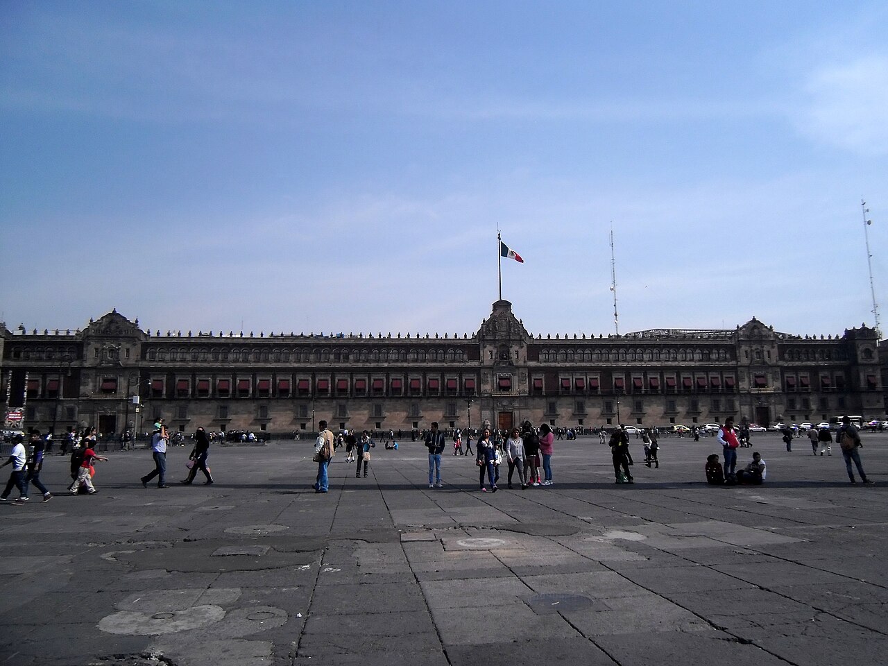 The long stone facade of the National Palace with a Mexican flag on top, seen across the Zócalo square with people walking