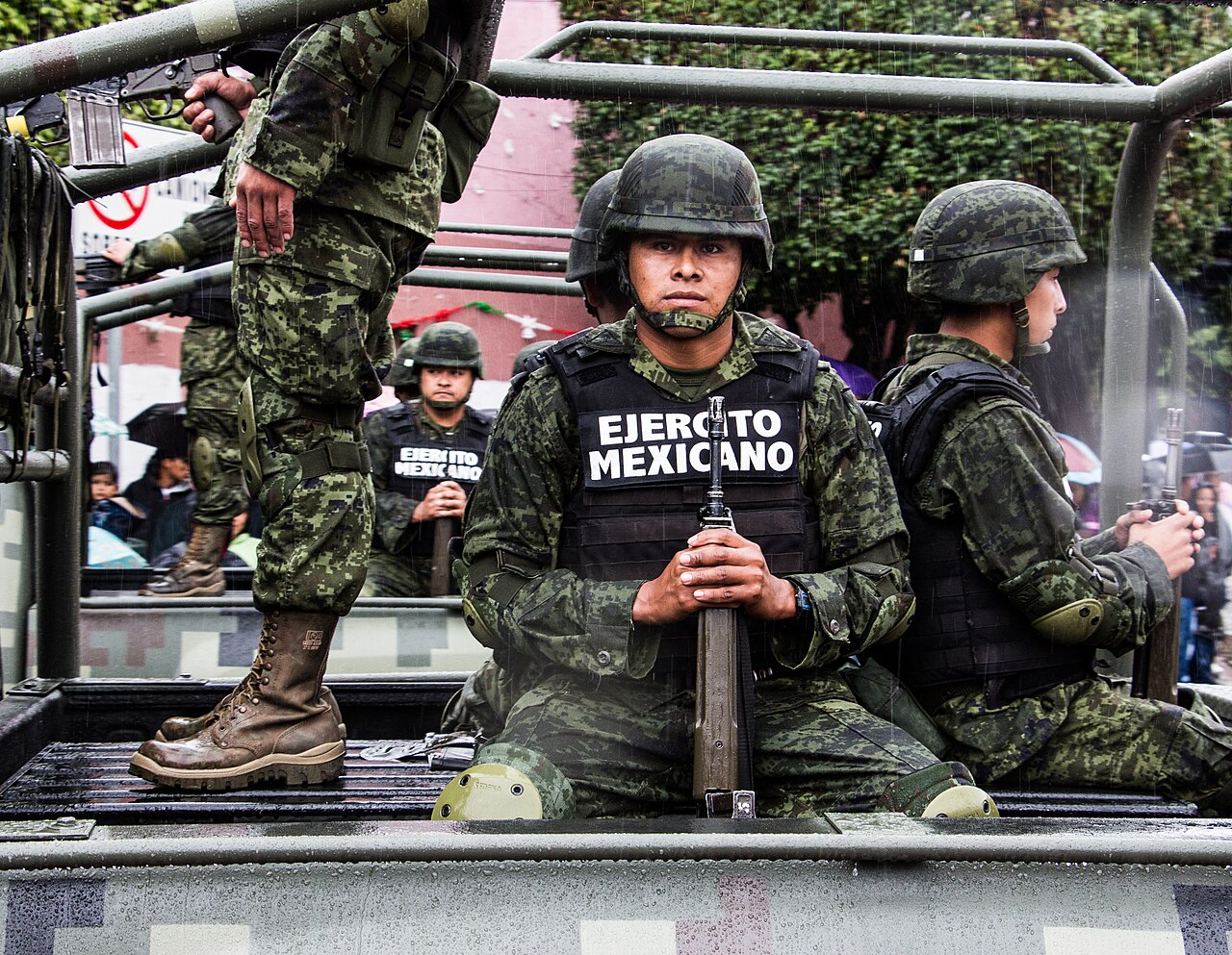 Mexican Army soldiers in helmets and vests marked Ejército Mexicano sit with rifles in the back of a military pickup truck in the rain