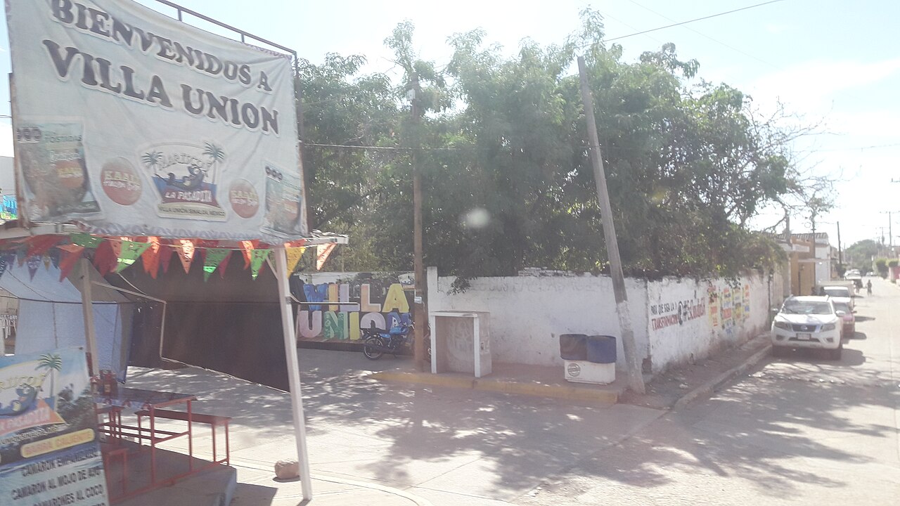 Street corner in Villa Unión, Sinaloa, with a Bienvenidos a Villa Union banner, a white wall with a colourful Villa Unión mural and parked pickup trucks