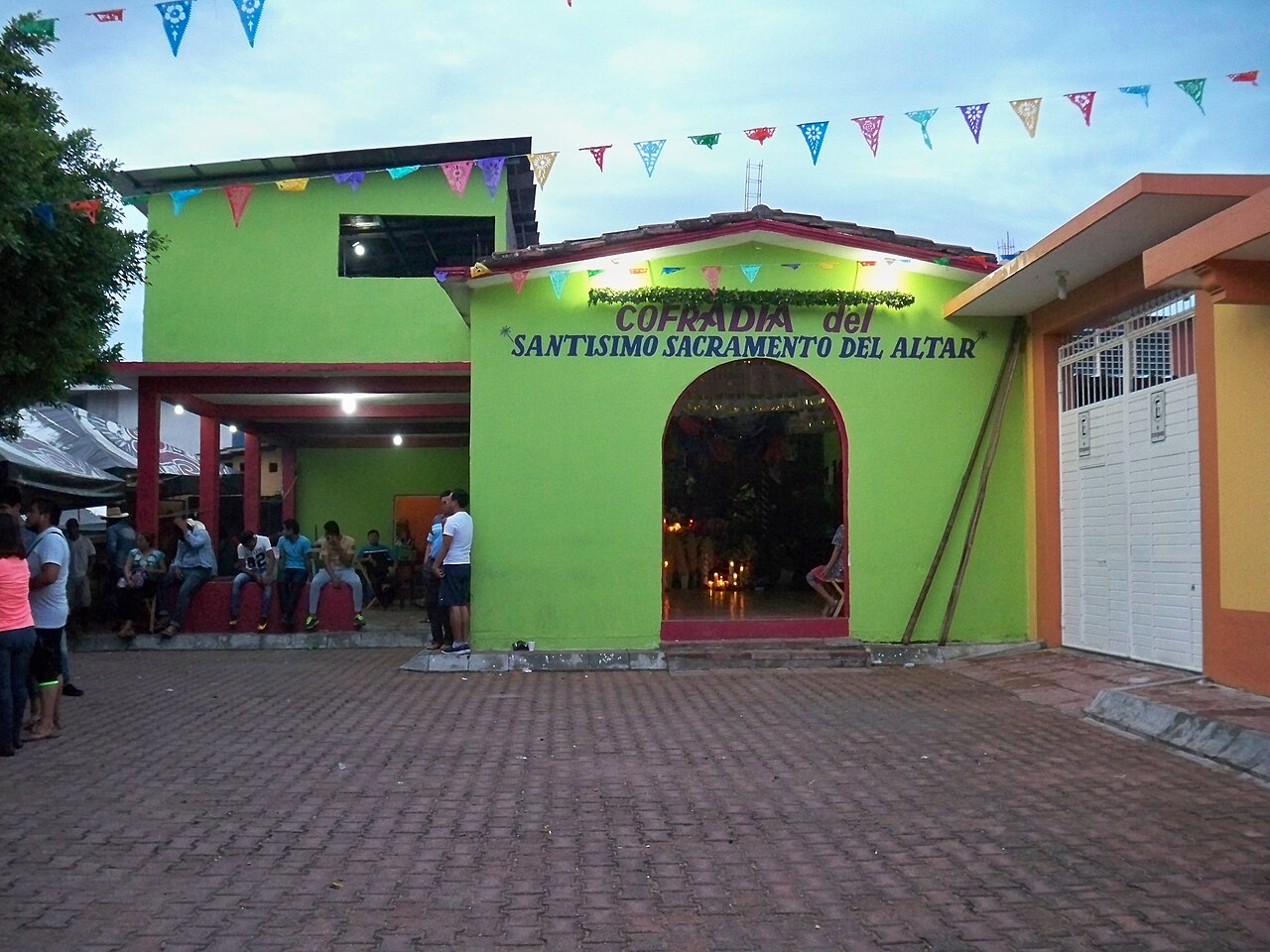 A lime-green brotherhood hall with an arched doorway on a brick-paved square in Suchiapa, Chiapas, with people sitting outside.