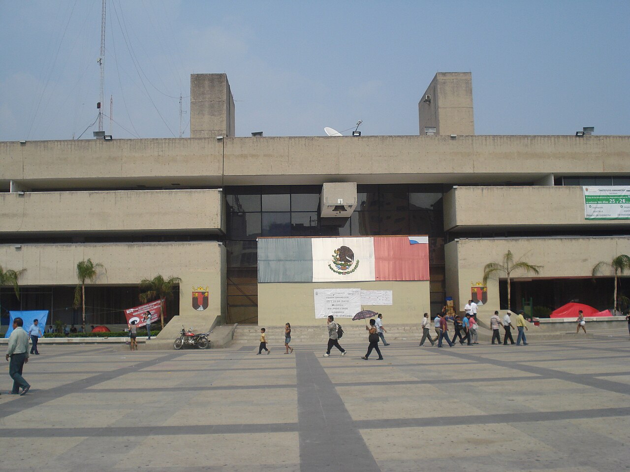 The concrete Chiapas Government Palace in Tuxtla Gutiérrez with a Mexican flag mural and people crossing the plaza in front.