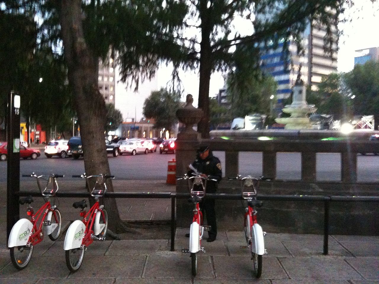 Red Ecobici bicycles docked at a station beside a fountain and evening traffic in Mexico City