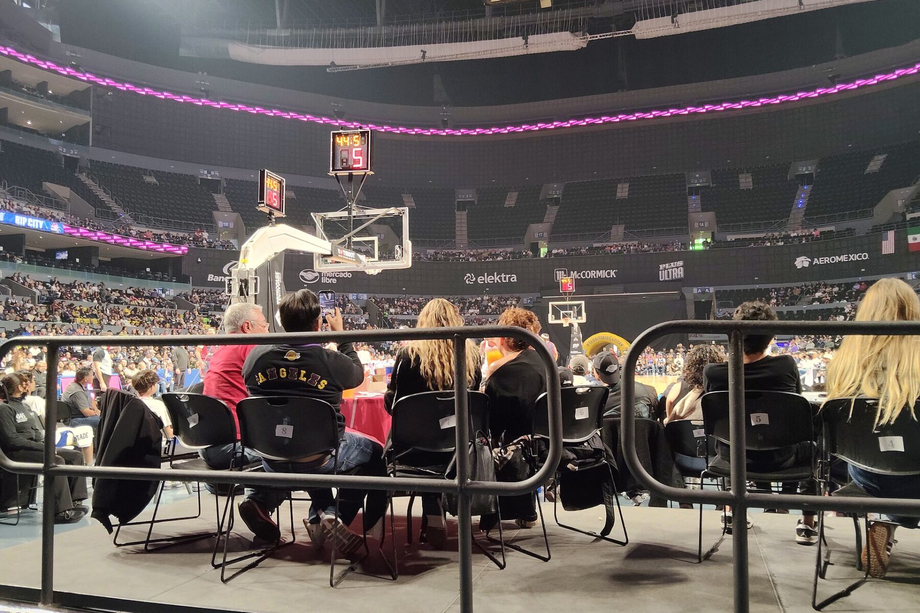 Fans watch a Mexico City Capitanes home game from courtside at Arena CDMX, with a pink LED ribbon board above the baskets and the lower and upper stands behind