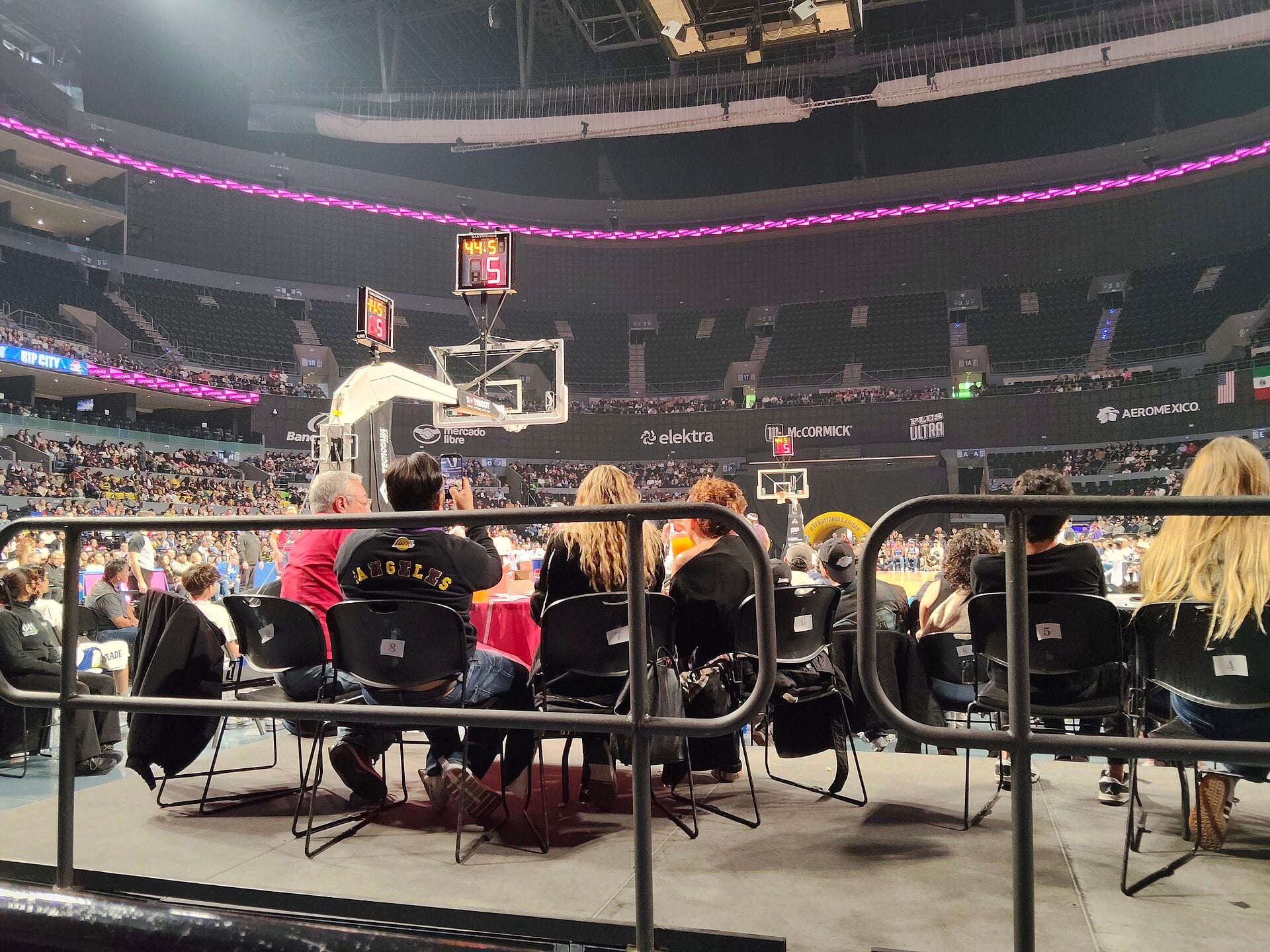 Fans watch a Mexico City Capitanes home game from courtside at Arena CDMX, with a pink LED ribbon board above the baskets and the lower and upper stands behind