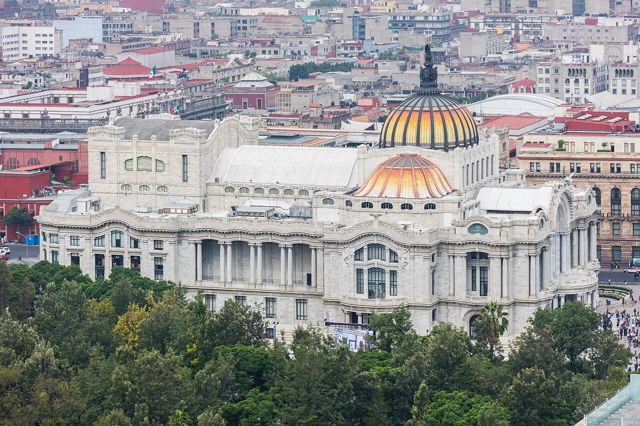 The marble Palacio de Bellas Artes with its coloured domes, seen from above among the rooftops of central Mexico City.