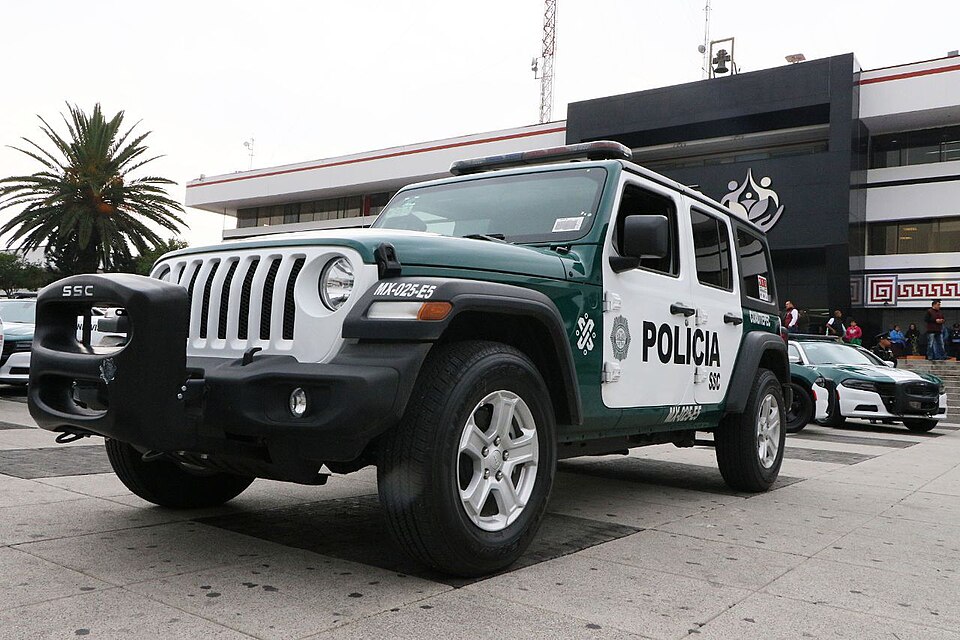 A green and white Jeep marked POLICÍA and SSC parked on a plaza in front of an office building, with more patrol cars behind it