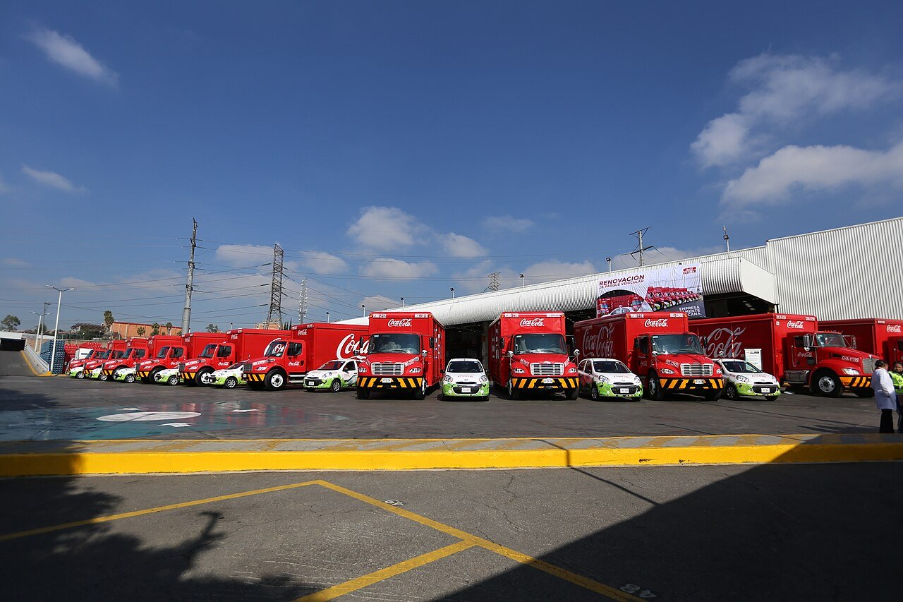 Red Coca-Cola delivery trucks and small cars lined up outside a Coca-Cola FEMSA distribution center in Mexico City