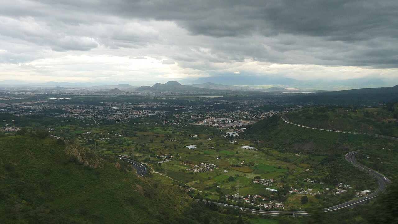 A highway curving through green hills above a wide valley of fields, villages and distant volcanic peaks under heavy cloud