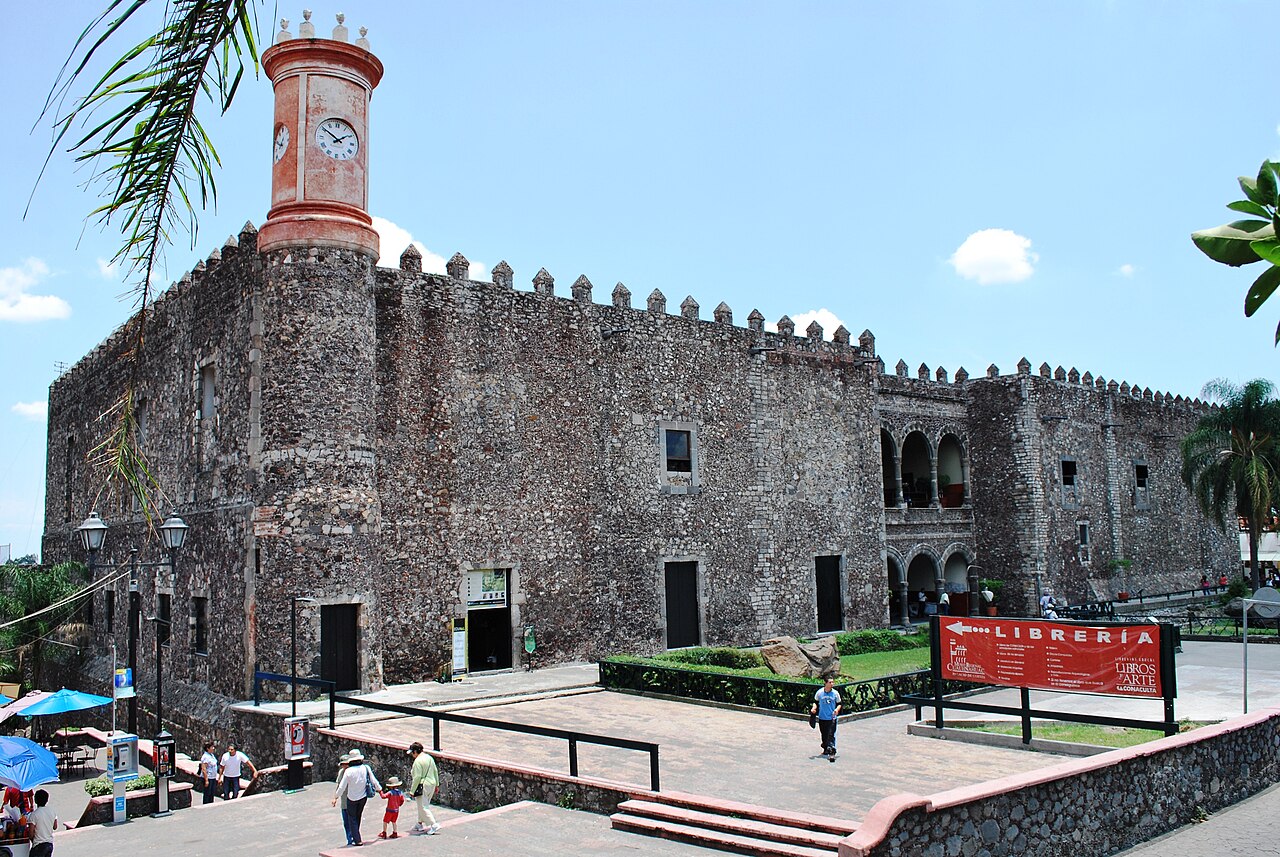 Visitors walk past the stone walls and clock tower of the Palacio de Cortés in Cuernavaca, Mexico, on a sunny day