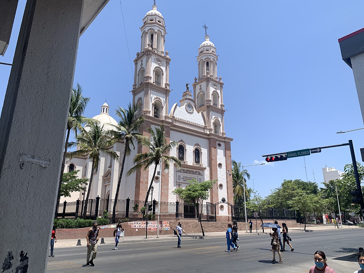 The twin-towered white and pink Cathedral of Culiacán beside a busy street with palm trees and pedestrians