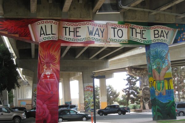 A colourful mural under a highway bridge in Chicano Park, San Diego, with a painted banner reading All the Way to the Bay.