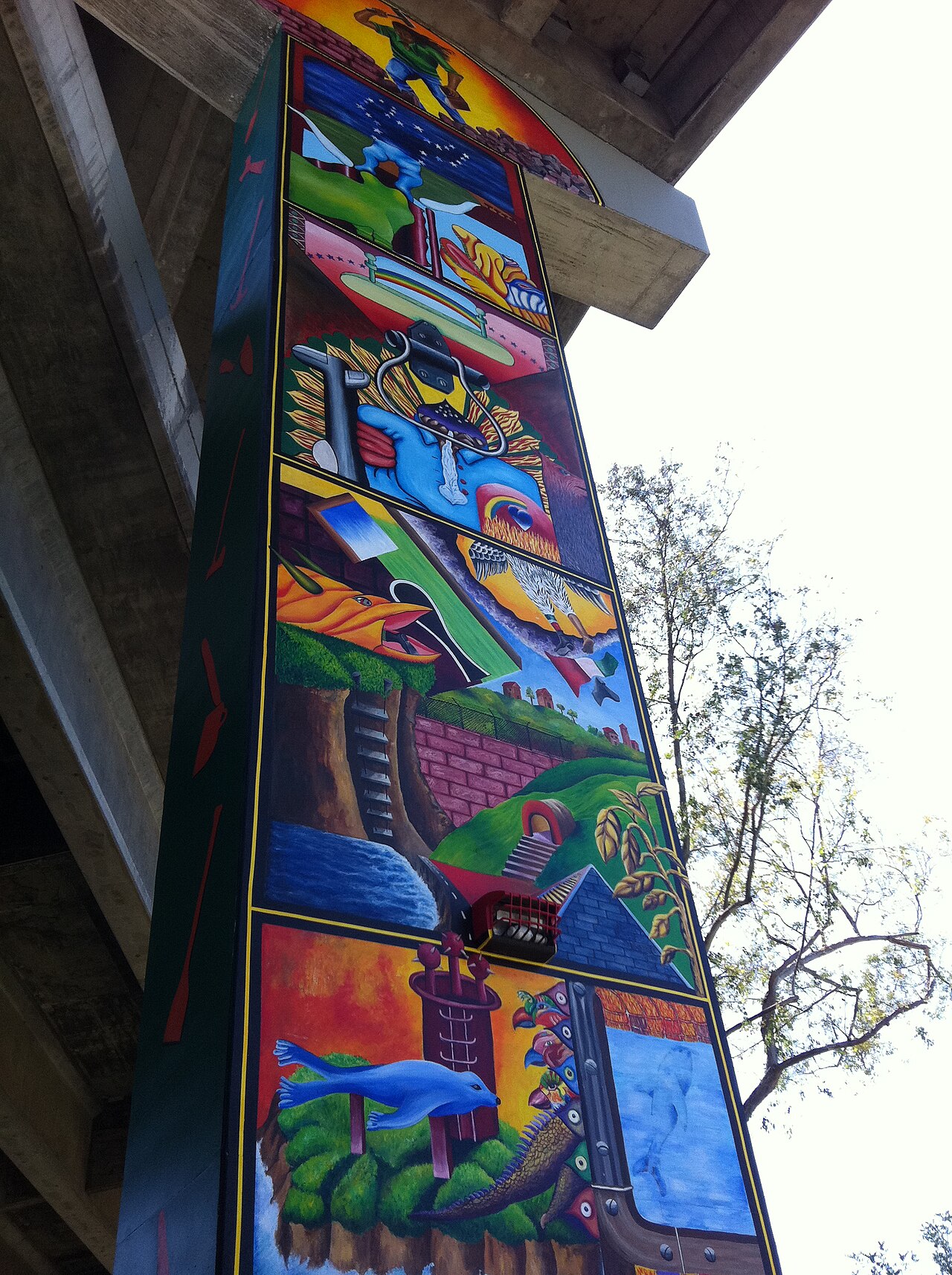 A tall painted pillar at Chicano Park in San Diego with colourful scenes of a worker, a border wall, a ladder and a pyramid.