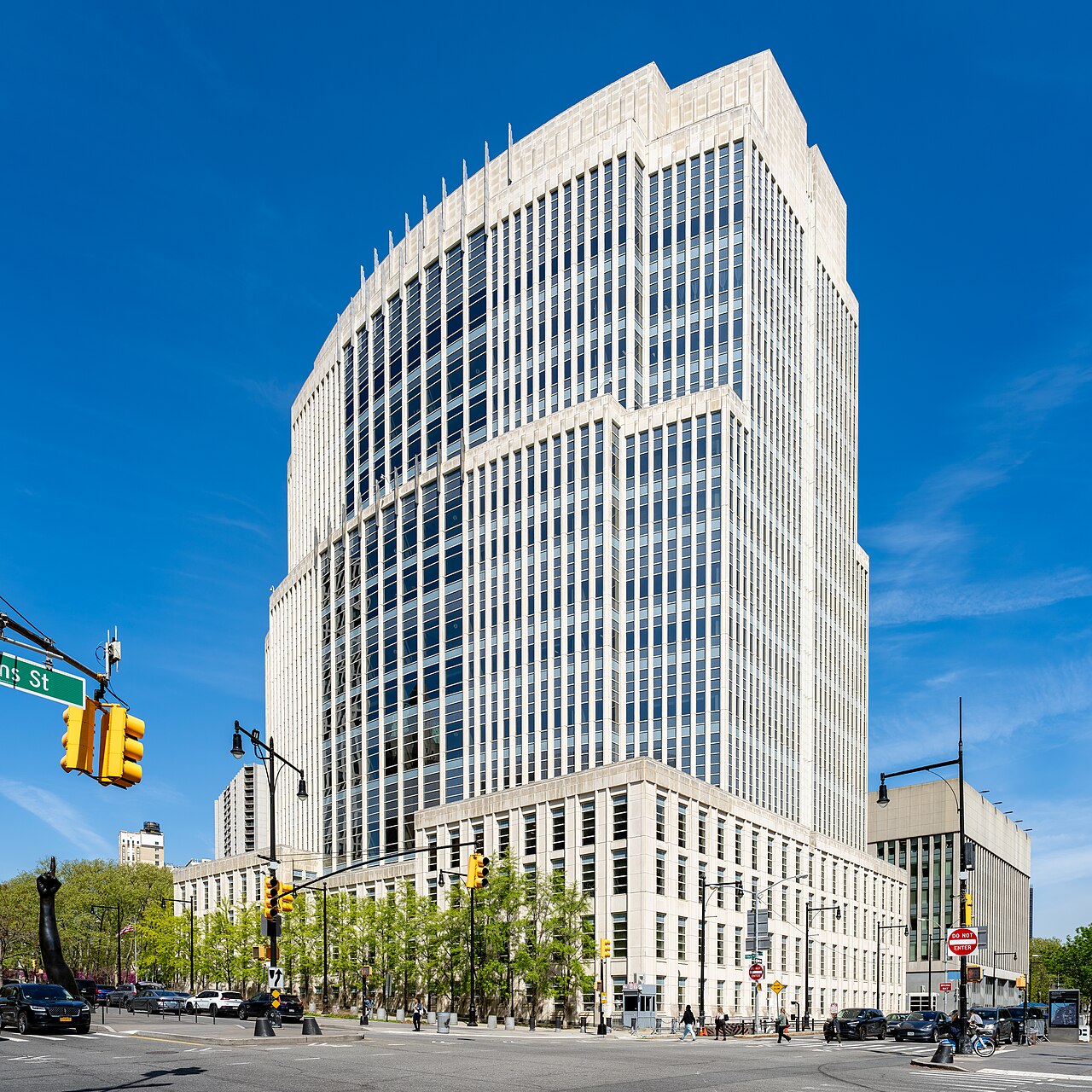 White stone and glass tower of the Theodore Roosevelt United States Courthouse in downtown Brooklyn under a blue sky, with traffic lights in the foreground