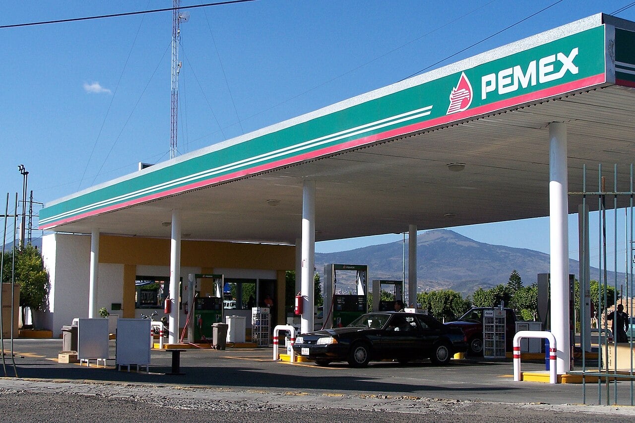 A Pemex filling station in Mexico under a green and white canopy, with cars at the fuel pumps and a mountain behind