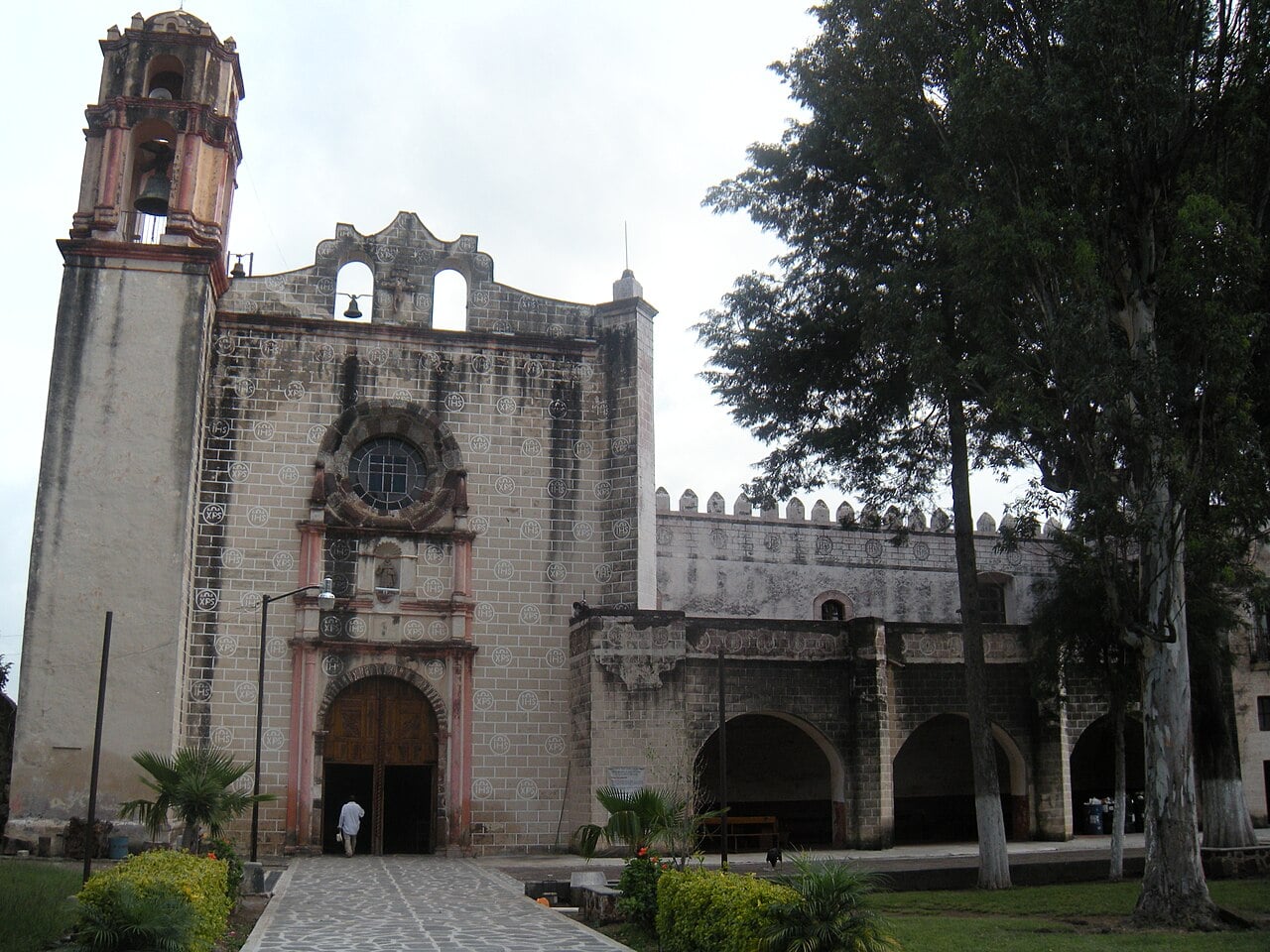 Stone facade and bell tower of the 16th-century San Guillermo convent church in Totolapan, Morelos, with trees and a paved path
